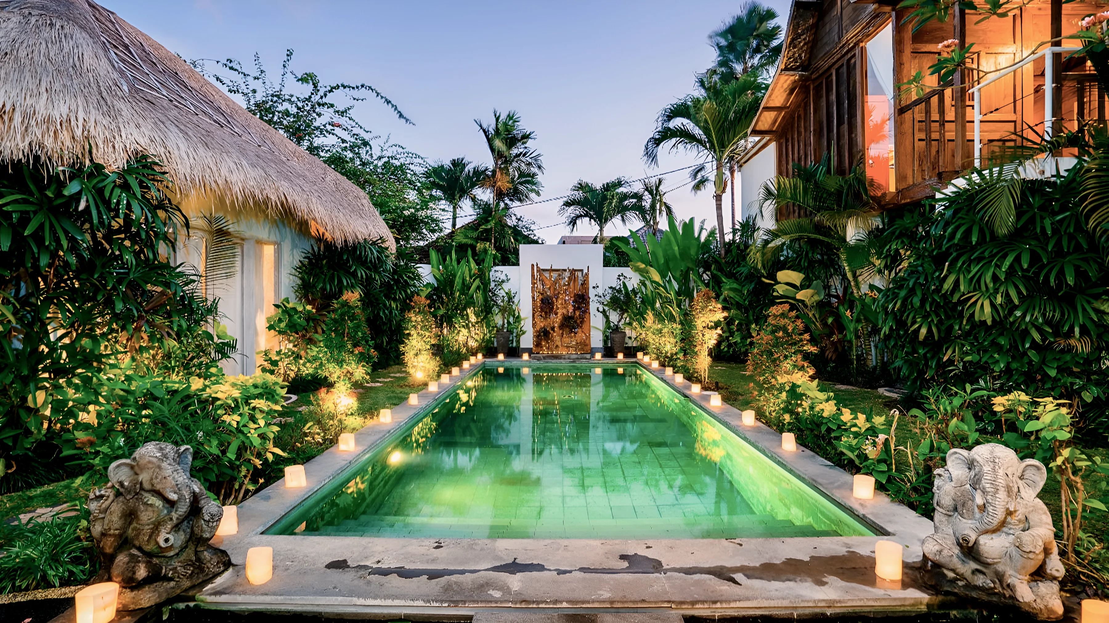 The pool lit at twilight, lanterns along both sides and the driftwood screen at the far end